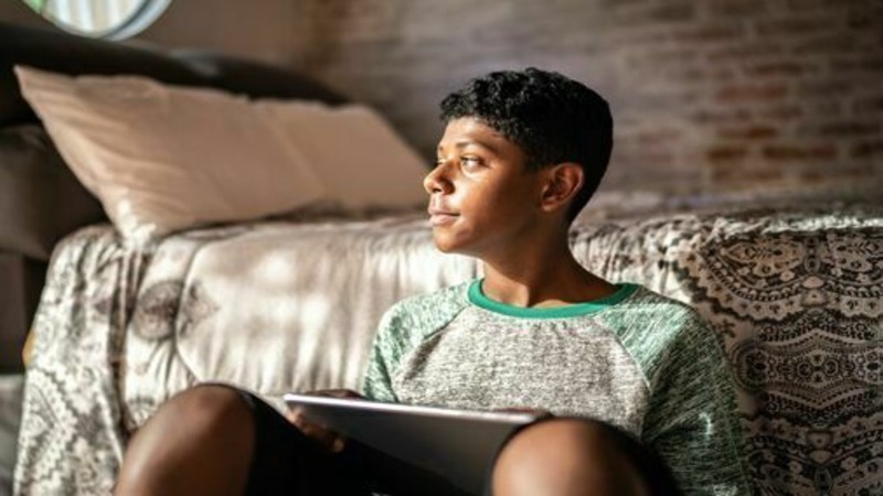 A boy with short curly hair sits on the floor, leaning against a bed, holding a tablet and looking to the left.